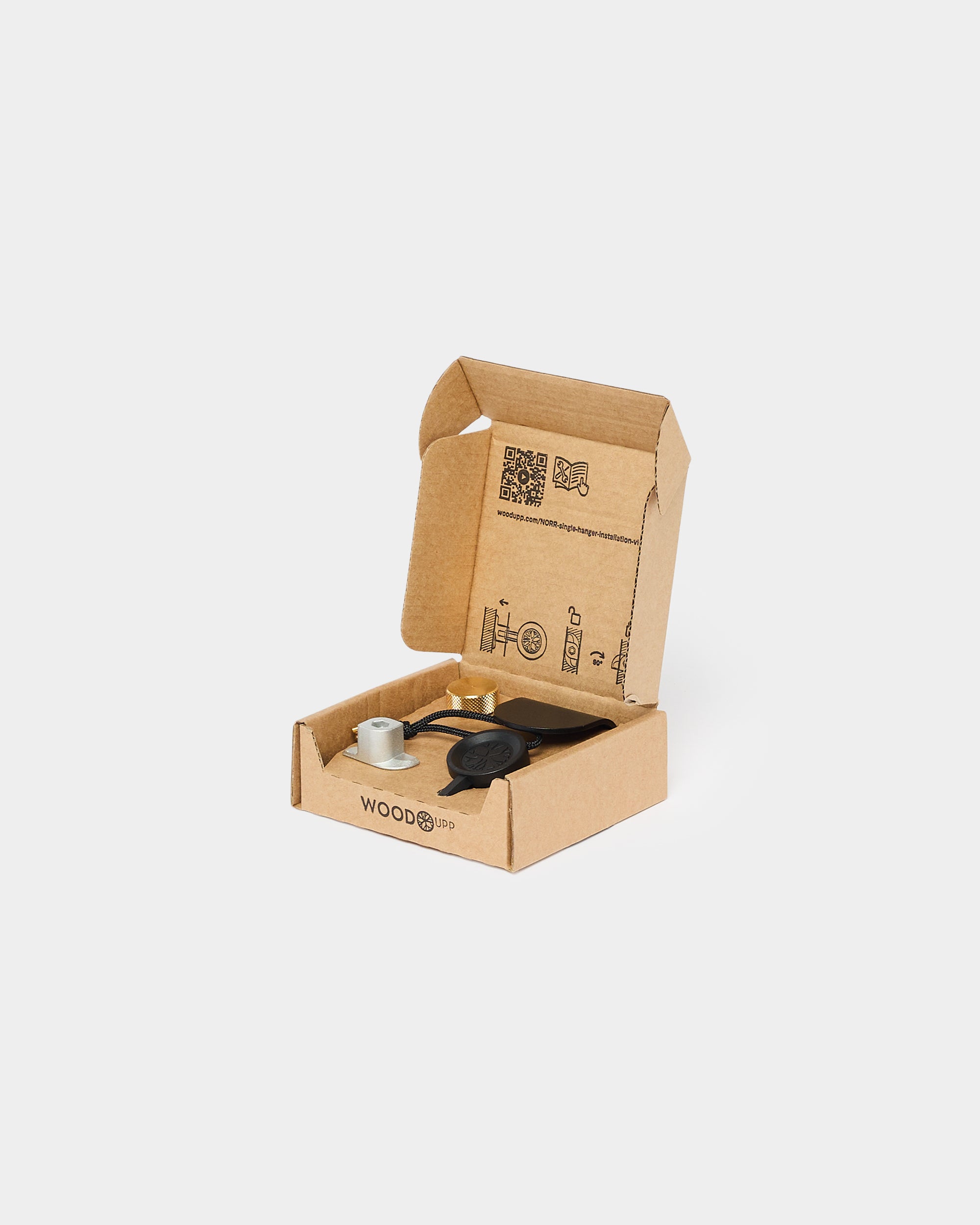 Small open WOODO cardboard box containing a round black acoustic accessory, a metallic fitting, and a gold pin, part of the WoodUpp CREATE collection.